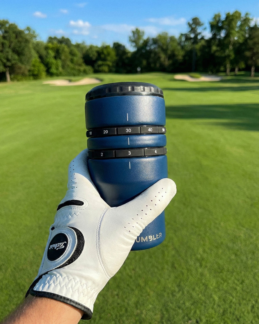 Blue tumbler with measurement markings held by a gloved hand on a golf course.